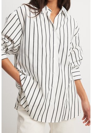 Kobieta ubrana w luźną białą koszulę na guziki z pionowymi czarnymi paskami i białe spodnie, jedna ręka w kieszeni, stoi na jednolitym tle.