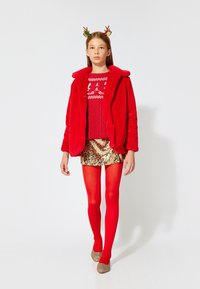 Rød faux fur-jakke over en rød striktrøje med festligt mønster, guld palietskørt, røde strømpebukser og guld sko. Antler-hårbåndet tilføjer detalje.