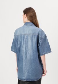 Τζιν πουκάμισο κοντομάνικο σε ανοιχτό μπλε χρώμα, με γιακά που κουμπώνει, άνετη εφαρμογή και ραφή στην πλάτη. Το ύφασμα έχει μια απαλή, ξεθωριασμένη υφή.