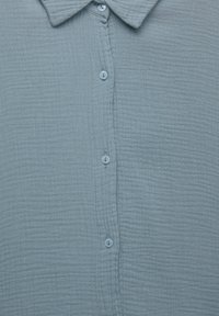 Chemise en tissu texturé bleu clair avec un col et cinq boutons visibles au milieu devant.