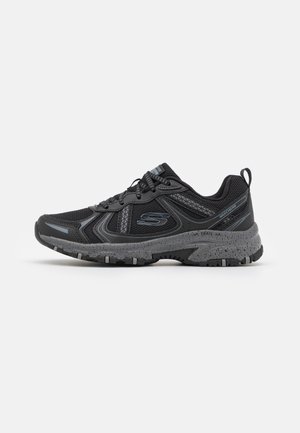 Chaussure de trail Skechers noire et grise avec tige en mesh, lacets, semelle texturée et languette au talon sur fond blanc.