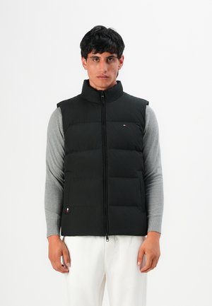 Man wearing black quilted sleeveless vest over gray long sleeves and white pants standing against white background.