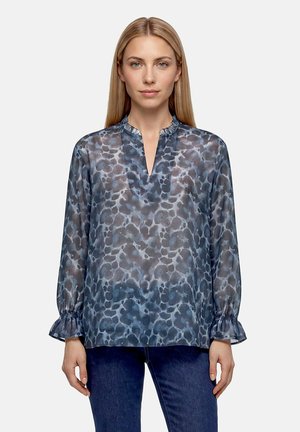 Frau trägt eine transparente, blau gemusterte Langarmbluse mit Rüschenkragen und Manschetten, kombiniert mit dunkelblauen Jeans, steht vor weißem Hintergrund.