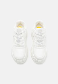 Zapatillas blancas con un material suave, puntera redonda y suelas gruesas. Presentan cordones prominentes y un acento amarillo en la lengüeta.