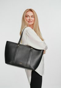 Czarna skórzana torba typu tote o teksturowanym wykończeniu, z złotymi akcentami i podwójnymi paskami na ramię. Prezentowana na jasnym tle.