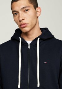 Sweat à capuche zippé bleu marine avec cordons blancs, comportant un petit logo sur la poitrine gauche et une texture de tissu agréable.