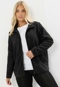 Svart fleecejacka med dragkedja framtill, ståkrage, långa ärmar och en slät textur. Bärs över en vit t-shirt och svarta leggings.