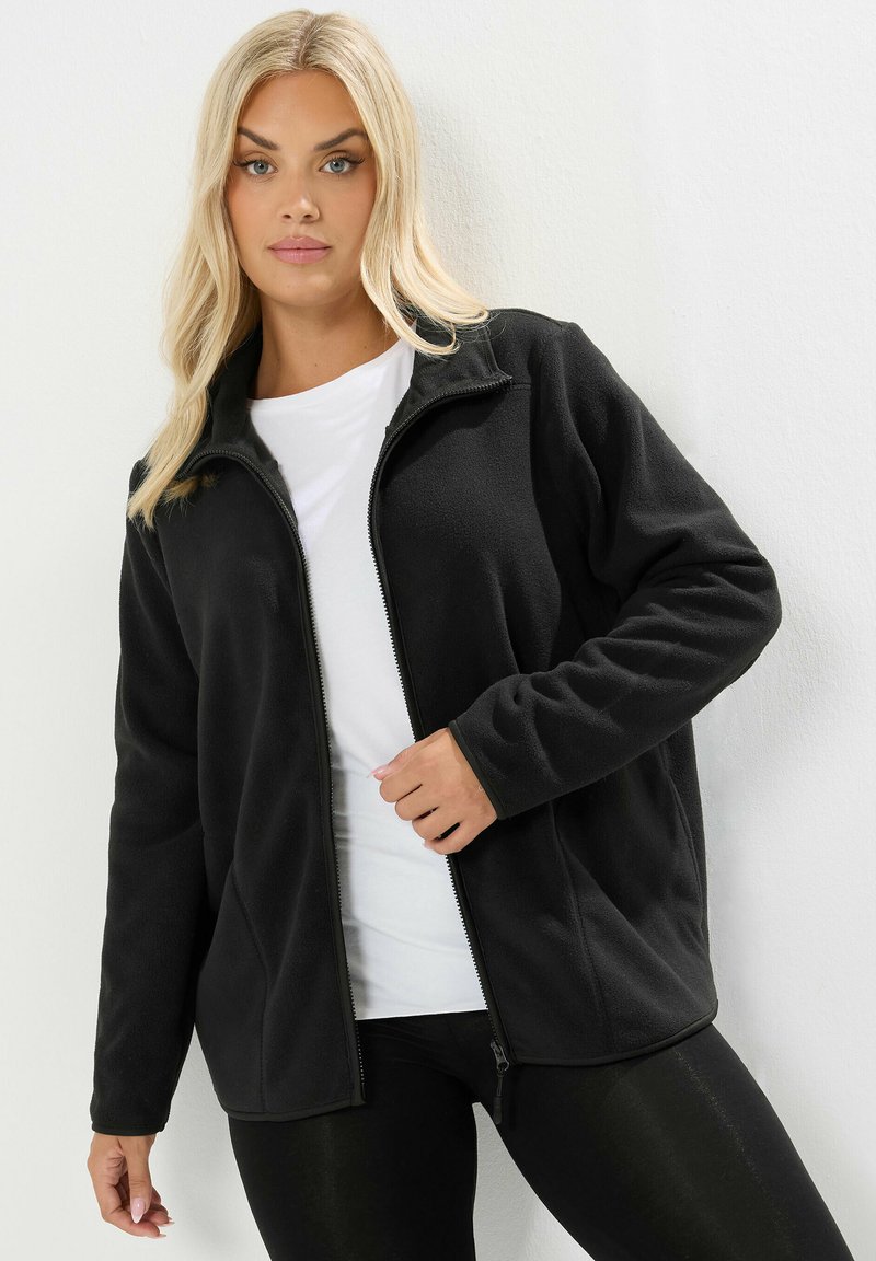 Svart fleecejacka med dragkedja framtill, ståkrage, långa ärmar och en slät textur. Bärs över en vit t-shirt och svarta leggings.