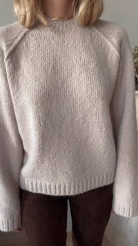 Light beige knit sweater with a textured finish, featuring a rounded neckline, raglan sleeves, and ribbed hem, paired with dark brown pants.