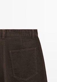 Pantalon en velours côtelé marron avec une texture nervurée, des passants pour ceinture et une poche arrière unique. Le tissu présente des rayures verticales et une couture solide.