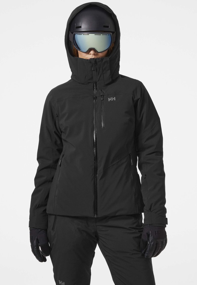 Schwarze Skijacke mit hohem Kragen, Reißverschluss vorne und Kapuze. Mit Reißverschlusstaschen und Logo auf der Brust. Kombiniert mit schwarzen Handschuhen und Hose.