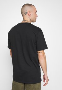 Camiseta de algodón negra con cuello redondo, mangas cortas y corte recto. Cuenta con una etiqueta en el dobladillo y tatuajes visibles en el brazo.