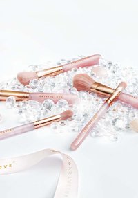 Zestaw różowych pędzli do makijażu z różowymi złotymi akcentami, z miękkim włosiem, ułożonych wśród przezroczystych dekoracyjnych kamieni i wstążki.