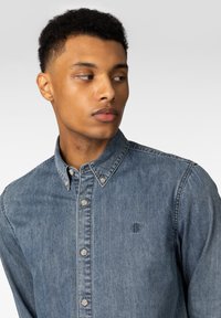 Denimskjorte i lyseblå, med krage, frontknapper og en liten logo på brystet. Myk tekstur, uformell design.