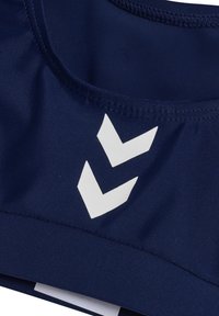 Granatowy top sportowy wykonany z gładkiego materiału, z białym logo w kształcie chevronu z przodu. Posiada okrągły dekolt i detale szycia.