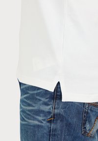 Camisa blanca de manga corta con aberturas laterales, combinada con jeans de mezclilla azules con textura deslavada y costuras en contraste.