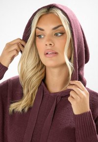 Bordeaux hoodie tillverkad av texturerad stickad tyg, med en justerbar huva och ribbade ärmmuddar, som framhäver en bekväm och avslappnad design.