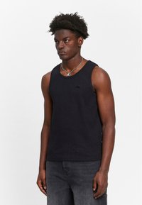 Black tank top made of cotton, featuring a fitted design, wide armholes, and a small logo detail on the chest. Worn with gray jeans.