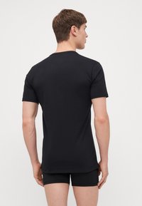 T-shirt en coton noir avec des manches courtes, un col rond et une texture lisse. Porté avec un short noir, montrant un design simple et minimal.