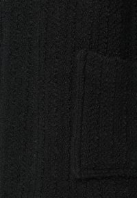 Zwarte gebreide jas met een patroon met textuur, voorzien van een opgestikte zak. Materiaal lijkt zacht met een licht pluizige afwerking.