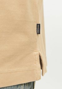 Beige színű pólo, texturált anyagból készült, oldalain hasítékkal a szegélyen, és egy kicsi fekete címkével, amelyen "JACK&JONES" felirat található az oldalán.