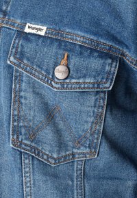 Nærbilde av en blå denimjakke-lomme med 'Wrangler'-merket og en sølvknapp med merkets logo.