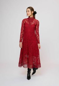 Červené midi šaty z čipky s dlhými rukávmi, vysokým golierom, priehľadnou vrstvou a viacvrstvovou sukňou. Nosia sa s čiernymi špicatými členkovými čižmami.