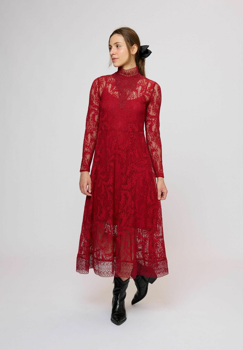 Červené midi šaty z čipky s dlhými rukávmi, vysokým golierom, priehľadnou vrstvou a viacvrstvovou sukňou. Nosia sa s čiernymi špicatými členkovými čižmami.