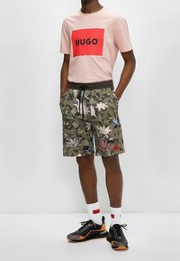 T-shirt rose clair avec un logo rectangulaire rouge, associé à un short kaki à motifs floraux et des baskets noires avec des accents orange.