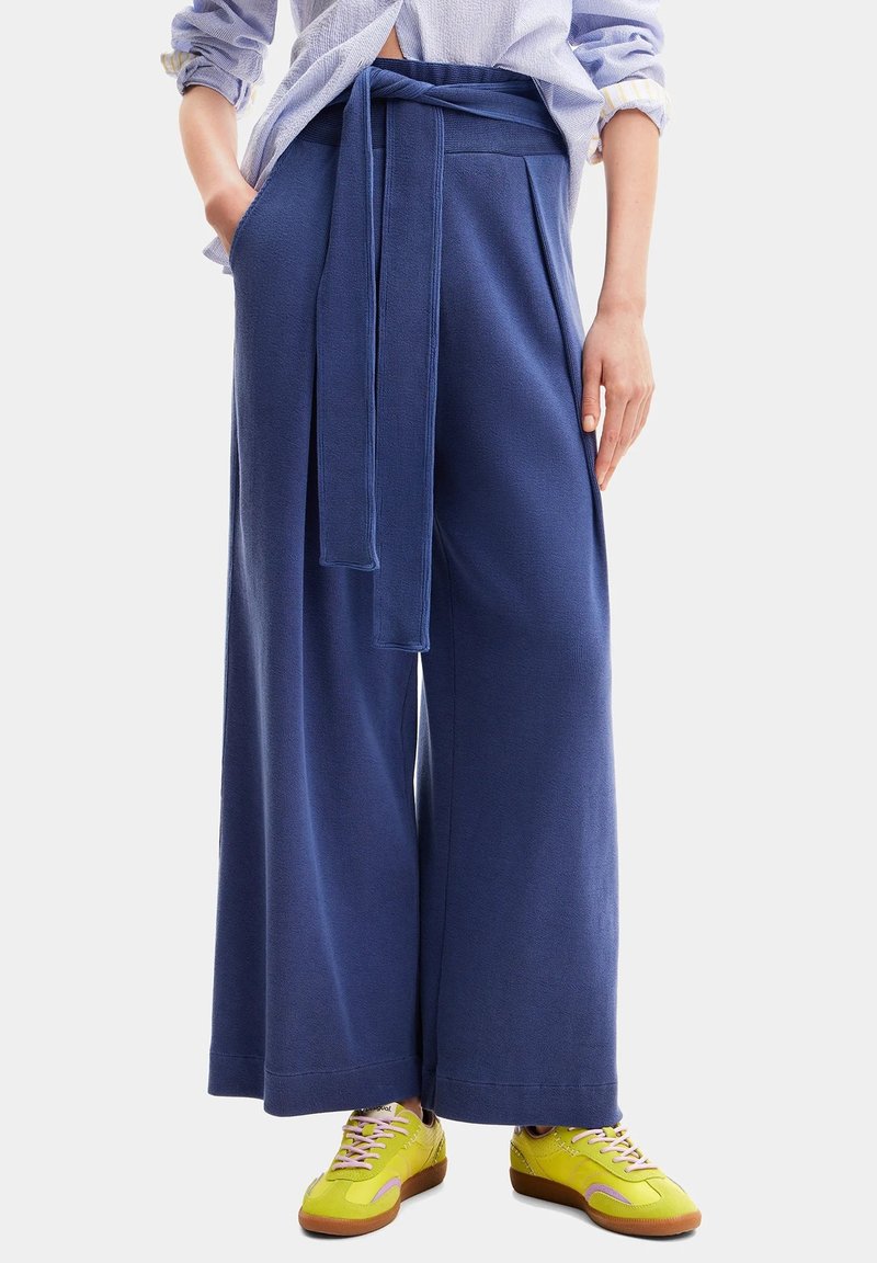 Pantaloni a gamba larga in tessuto blu navy con una finitura testurizzata, dotati di una vita con laccetto e tasche laterali. Abbinati a sneakers gialle.