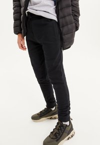 Pantalon de survêtement noir à coupe fuselée, texture douce, taille élastique, poches latérales, associé à des baskets vertes et noires avec des semelles volumineuses.