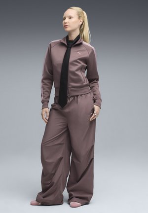 Ensemble de sport en tissu mauve, comprenant une veste zippée avec cordon noir et un pantalon à jambes larges, texture lisse, avec un détail de logo sur la veste.