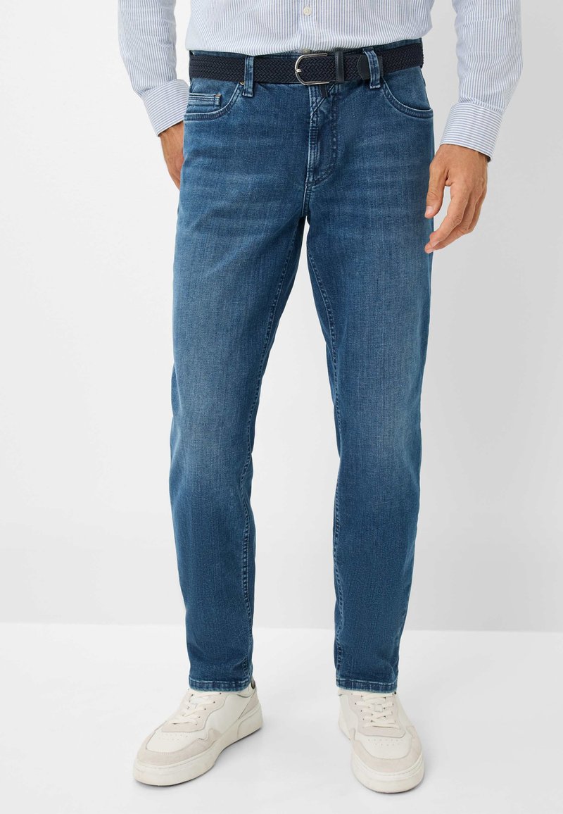 Jeans in denim blu con una vestibilità dritta. Caratteristiche includono una cintura scura e scarpe da ginnastica bianche. Il materiale appare morbido con una leggera texture lavata.