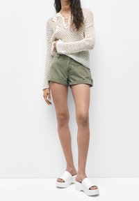 Top blanco de malla de manga larga con un agujero en el cuello, shorts enrollados de color verde oliva y sandalias de plataforma blancas. Texturas suaves; diseño casual.