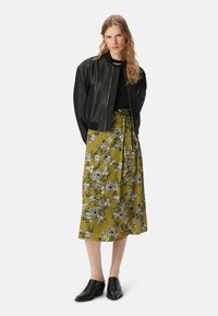 Giacca bomber nera in pelle sopra un top nero, abbinata a una lunga gonna floreale verde oliva. Le scarpe nere con tacco largo completano l'outfit.