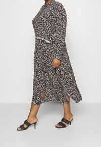 Vestido negro con estampado floral y mangas largas, con aberturas laterales y cinturilla blanca de marca. Combinado con sandalias de tacón negro con tiras.