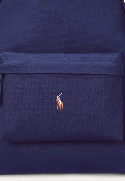 Tuvplāns tīkamas auduma mugursomas kabatā ar nelielu izšūtu polo spēlētāju, kurš valkā sarkanu cepuri un jāj zirga mugurā.