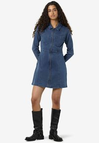 Denimdress med krage, lange ermer, to frontlommer og glidelås foran. I blått med synlige sømdetaljer.