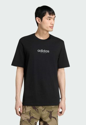 Młody mężczyzna w czarnej koszulce Adidas z logo i spodniach maskujących, stoi na jednolitym jasnym tle, patrzy w prawo.