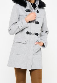 Žena oblečená ve světle šedém vlněném kabátu s černým kožíškem na límci, třemi černými knoflíkovými zapínáními, kapsami na klopě a hnědými kalhotami, ruka v kapse.