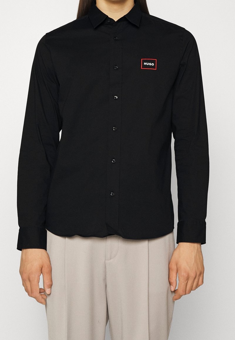 Homme portant une chemise noire boutonnée avec un petit logo "HUGO" bordé de rouge sur la poitrine gauche et un pantalon beige clair.