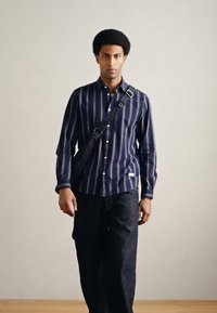 Koszula z guzikami w granatowe paski z białymi akcentami, zestawiona z ciemnymi spodniami dżinsowymi. Przez tors przechodzi czarna torba na pasku na ramię.