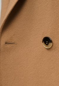 Ύφασμα παλτού σε ταμπά χρώμα με διακριτή υφή, που διαθέτει ένα μεγάλο, σκούρο κουμπί και μια μικρή τσέπη με φερμουάρ. Καθαρές γραμμές και ραμμένο σχέδιο.