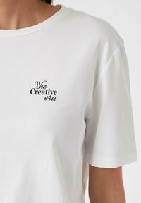 Weißes Baumwoll-T-Shirt mit kurzen Ärmeln und Rundhalsausschnitt; verfügt über schwarzen gestickten Text "The Creative era" auf der Vorderseite.