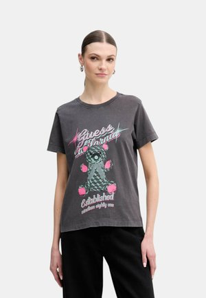 Kobieta o ciemnych włosach związanych w niski kucyk, ubrana w grafitowy T-shirt z różowo-miętowym nadrukiem misia i napisem "Guess California" oraz czarne spodnie.