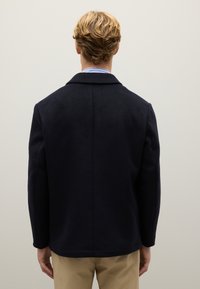 Una persona bionda, vestita con una giacca blu scuro e pantaloni beige, si trova di fronte a un muro grigio chiaro e uniforme.