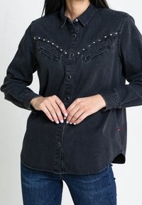 Black denim shirt featuring snap studs along the shoulders and two front pockets. Buttons down the front, and a relaxed fit with a rounded hem.