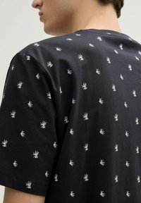 Camiseta negra de manga corta con patrones de hojas de palma bordados en blanco. Tela de algodón suave con un corte relajado y un cuello redondo.