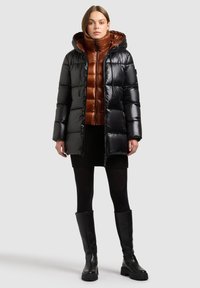 Svart vadderad jacka med huva, glänsande yta och figursydd design. Bärs över ett brunt underställ, båda kombinerade med en svart kjol.