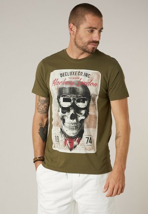 Homme portant un T-shirt vert olive avec un grand motif de crâne portant des lunettes de protection et une écharpe rouge, associé à un pantalon blanc, debout les mains dans les poches.